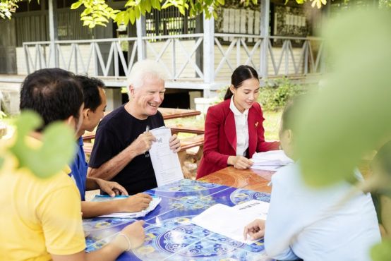 A group of people sit around a table with papers and pens on the table in front of them. A man holds up a piece of paper so it's visible to others at the table, as though instructing or teaching. He is laughing or smiling candidly, and others at the table are either watching on, or looking their own papers.