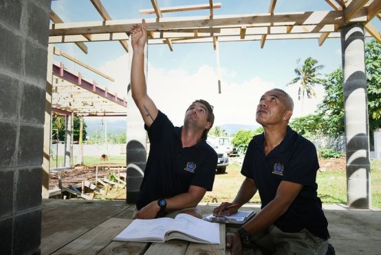 Two men wearing navy blue polo shirts crouch in a building site. The man on the left points directly above him, and the man on the right follows his line of sight. A book such as a technical manual or similar is open on a flat surface in front of them.