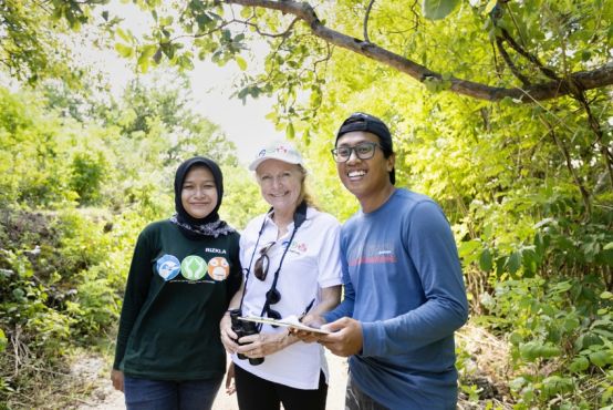 Three people stand shoulder-to-shoulder in an outdoor area with lots of trees in the background. They are smiling into the camera, and the person in the middle wears binoculars around her neck.