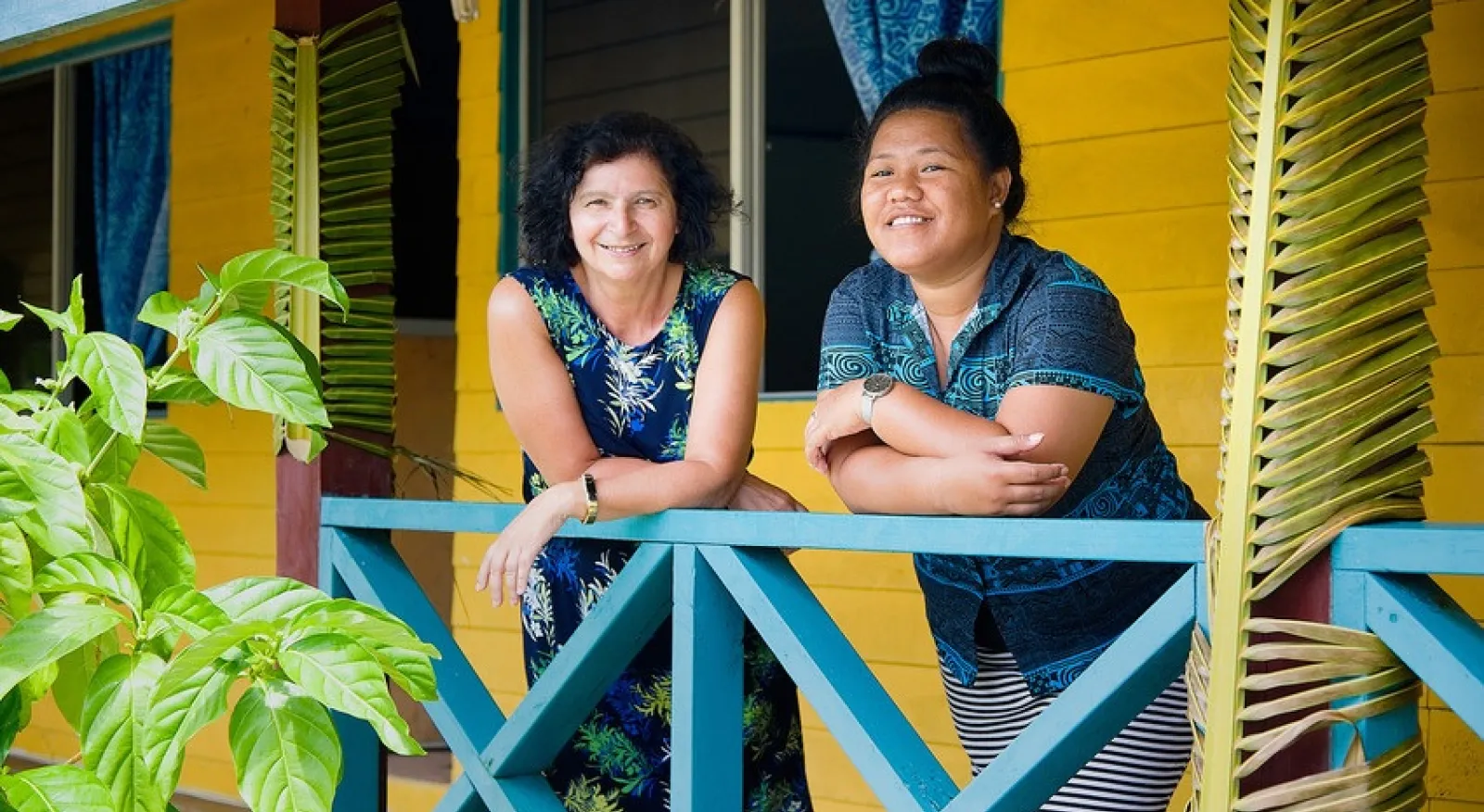 Two women stand on the veranda of a brightly coloured weatherboard building, leaning their elbows on the railing and smiling directly into the camera.