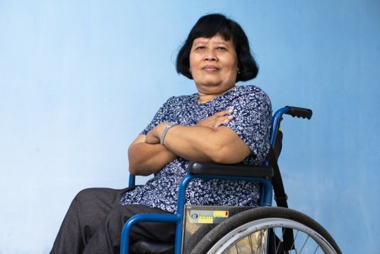A woman looks directly into the camera. She is in a wheelchair, with a pastel blue wall behind her. Her arms are crossed in a positive and steadfast expression.