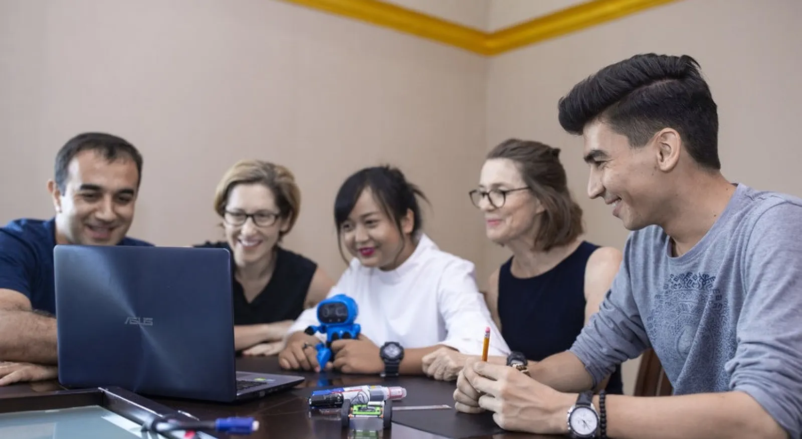 Five people sit at a table, all looking at a laptop and smiling. On the table near the laptop are various small gadgets, and one of the people holds a small robot in her hands.