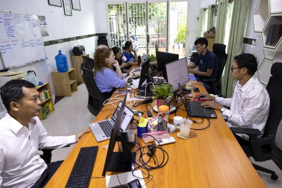 Six people all sit around a very large table, each with their own set up of computer screens and desk bric-a-brac in front of them. There are four people on the left side of the table, and two on the right. They are captured candidly working, each looking at their screens.