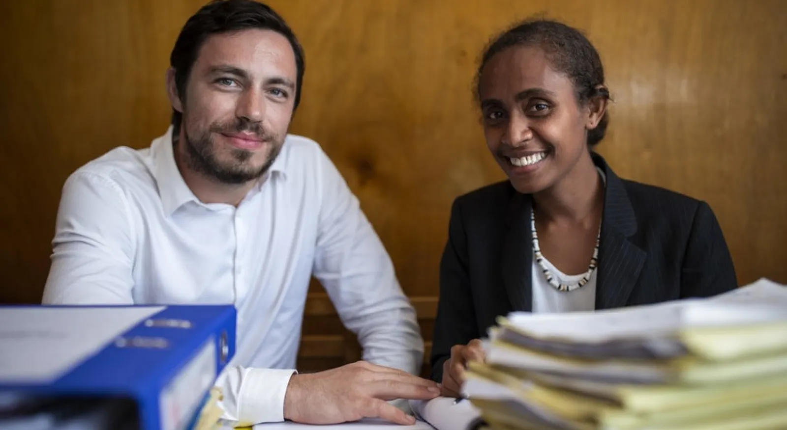 A man and a woman wearing neat business attire are seated at a table which is piled high with files and papers. They both smile directly into the camera.