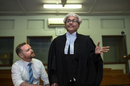 In a court room, a man is seated wearing a collared business shirt and tie. He looks up at a person standing beside him, who is wearing a legal wig and robes.
