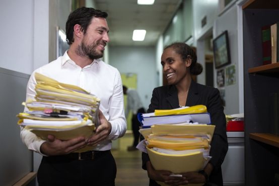 Two people walk down a corridor, each carrying a large stack of papers and files in their arms. They are smiling candidly at each other, as though in conversation.