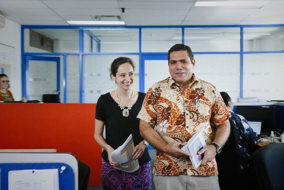 A woman and a man walk through an office environment; the woman is smiling and the man wears a candid, neutral facial expression.