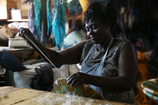 A woman sits at a table smiling candidly as she uses both her hands to weave a basket with an elaborate design.