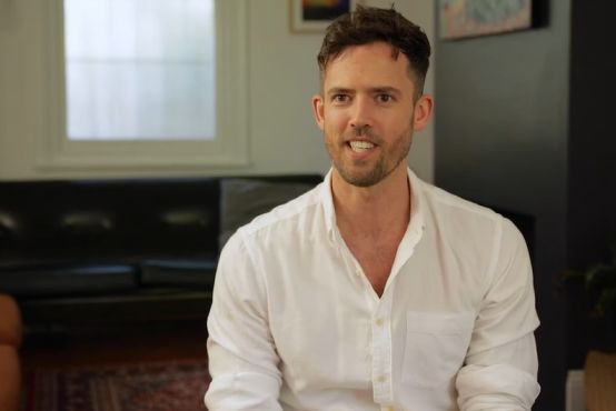 A man wearing a white collared shirt sits slightly to the right in the frame, looking candidly into the camera.