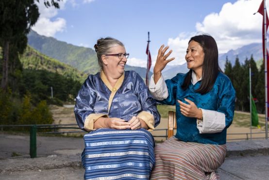 Two women sit outdoors with mountains, trees and a cloudy blue sky in the background. In the foreground, the women are chatting in sign language.