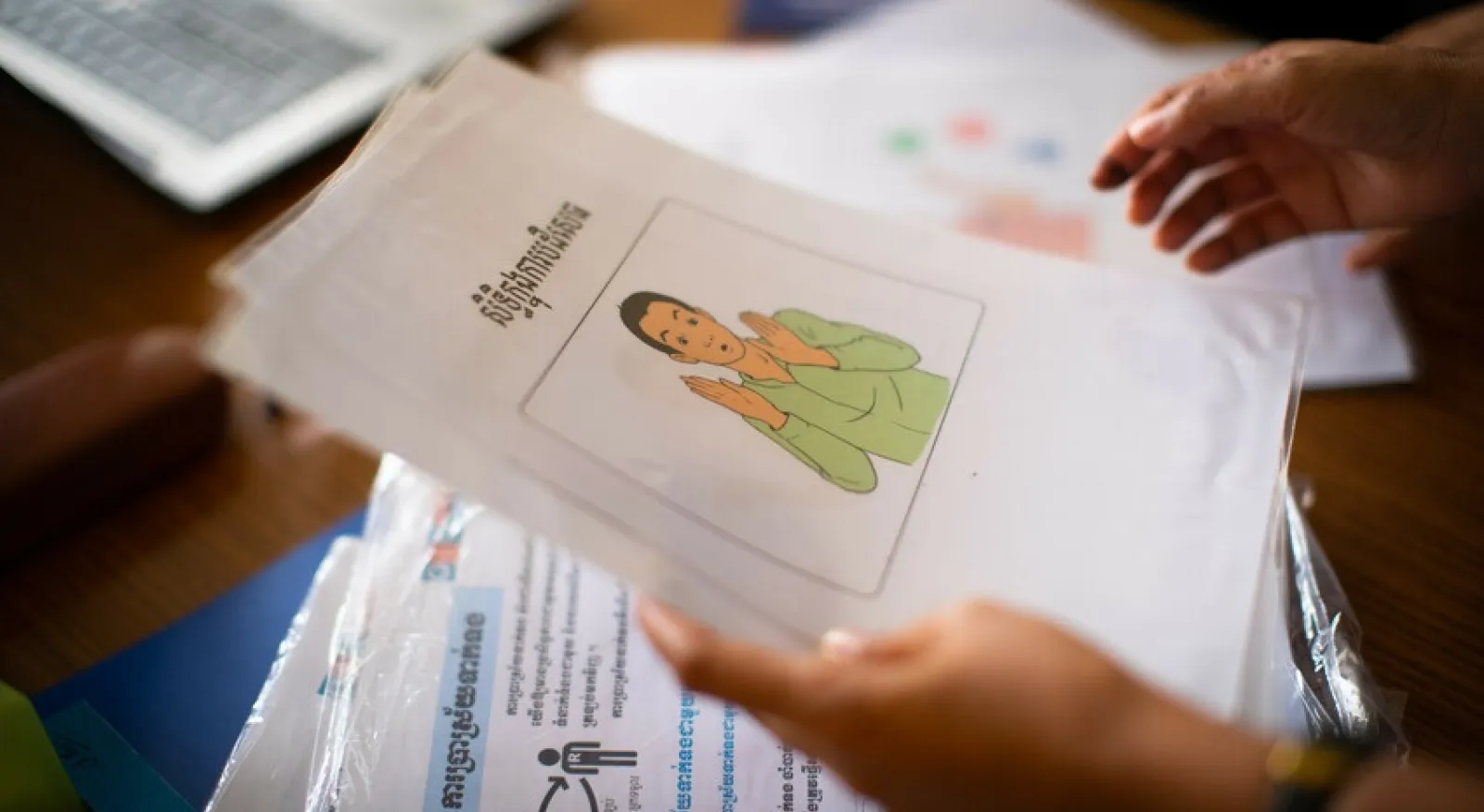 Hands holding a piece of paper with a picture of a person. There is writing in Khmer and the person is making hand gestures.