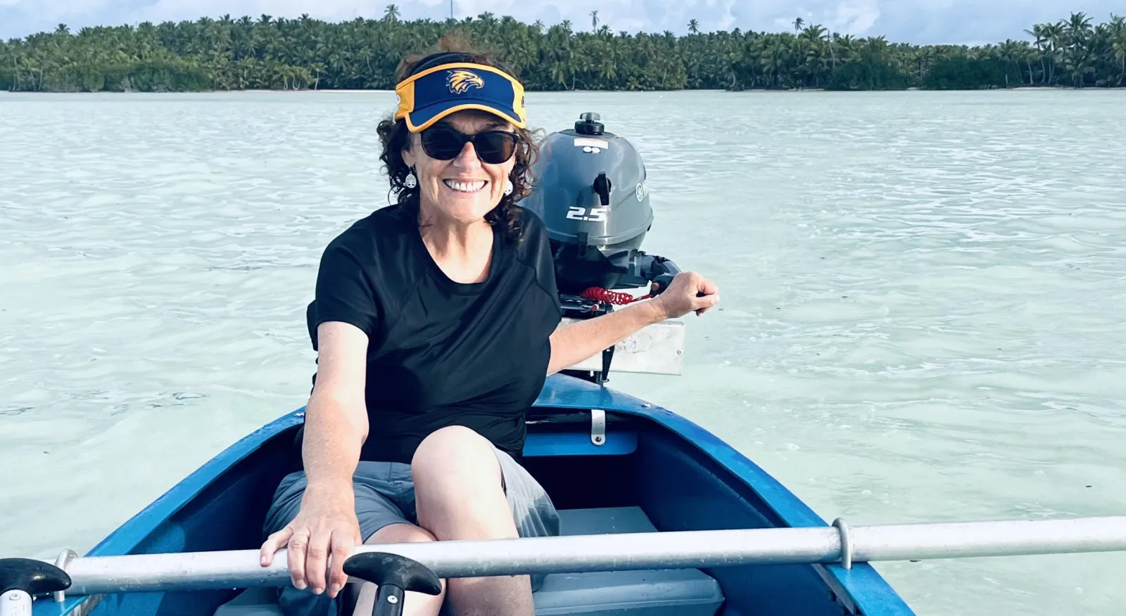 A woman smiles into the camera, sitting in a blue dinghy. She has one hand on the steering arm of the engine, and another hand on a horizontal metal bar in the boat. She is wearing a navy t-shirt, grey shorts, sunglasses and a sun visor.