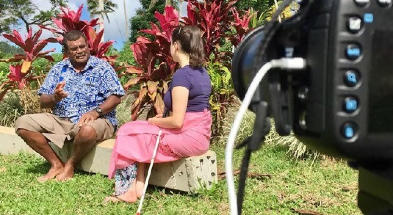 Two people sitting on a bench talking. The man is explaining something to the woman, who has a white cane by her side. They are being filmed by a camera.