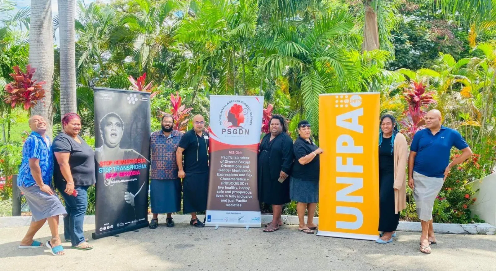 A group of eight people standing in a line outdoors with lush greenery behind them, and three banners reading: 'Stop transphobia'; 'Pacific sexual and gender diversity network'; and 'UNFPA'