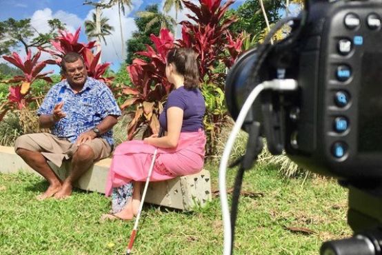 Two people sitting on a bench talking. The man is explaining something to the woman, who has a white cane by her side. They are being filmed by a camera.