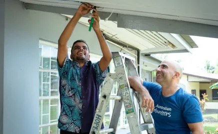 A person stands smiles as they stand on the lower rungs of a ladder with both arms raised above their head. They use a hand tool to fix something in a roof. A second person stands on the other side of the ladder smiling and watching on.