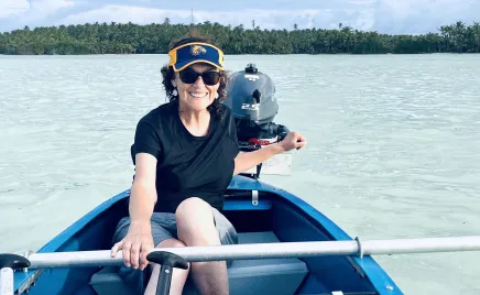 A woman smiles into the camera, sitting in a blue dinghy. She has one hand on the steering arm of the engine, and another hand on a horizontal metal bar in the boat. She is wearing a navy t-shirt, grey shorts, sunglasses and a sun visor.
