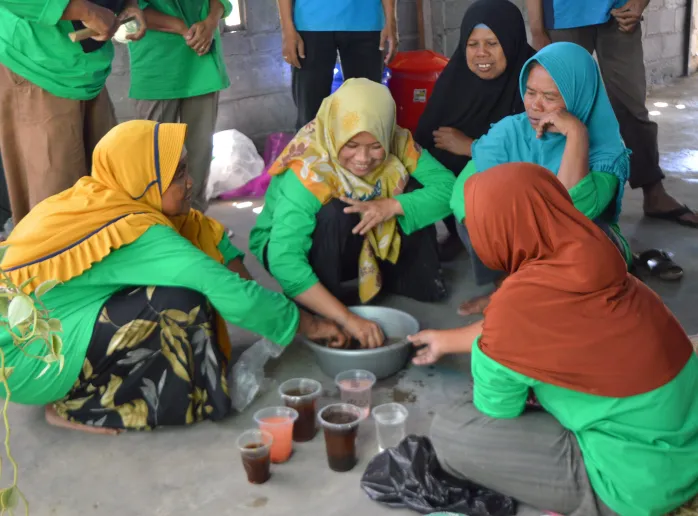 Five women are sitting on the ground working with soil and compost in a small bowl. They are smiling and all are wearing hijabs.