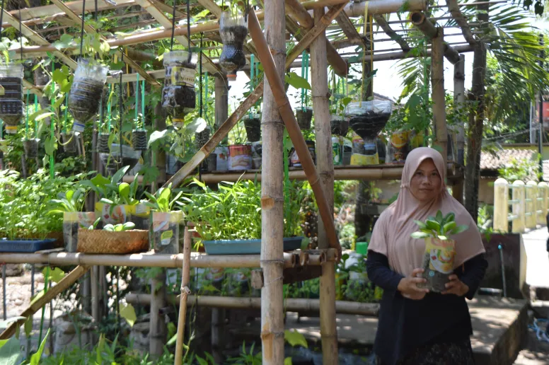 A woman is standing in an outdoor garden filled with plastic bottles and other containers which have seedlings and other plants growing.