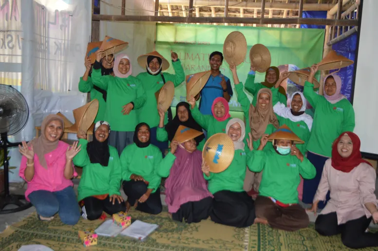A group of 16 women looking at the camera and smiling. Some are holding up traditional hats. Most are in green PPMK shirts.