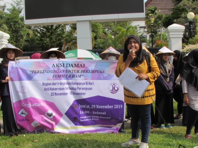 A group of women protesting and holding a larger poster that says, "Peaceful Action - Protection for women's rights defender". One women stands at the front holding a microphone.