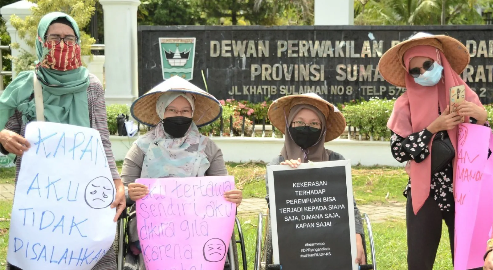 Four women stand in front of an Indonesian government building, holding signs calling for womens rights. Two women are in wheelchairs, and their friends stand next to them. All four women wear headscarves and masks, and look cheerfully at the camera.