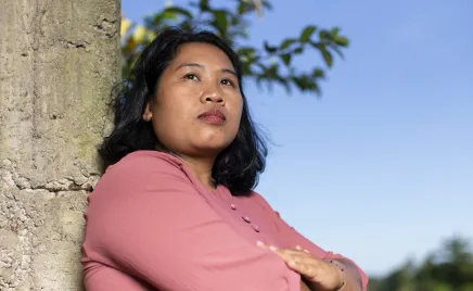 A woman is folding her arms and looking into the distance with a serious expression. She is leaning against a tree with a blue sky backdrop. She has a pink shirt and shoulder length black hair.