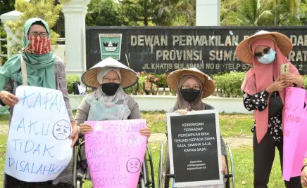 Four women stand in front of an Indonesian government building, holding signs calling for womens rights. Two women are in wheelchairs, and their friends stand next to them. All four women wear headscarves and masks, and look cheerfully at the camera.