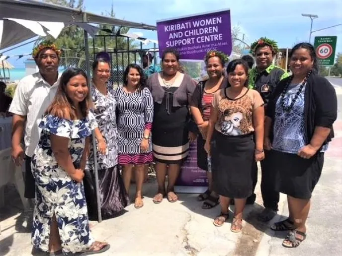A group of nine people standing and smiling infront of a sign that reads, "Kiribati women and children support centre"