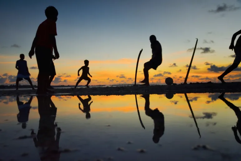 The silhouettes of people playing soccer on a beach against a sunset.