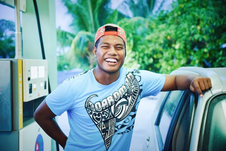a person with a backwards cap and t-shirt leans against a car, smiling widely.