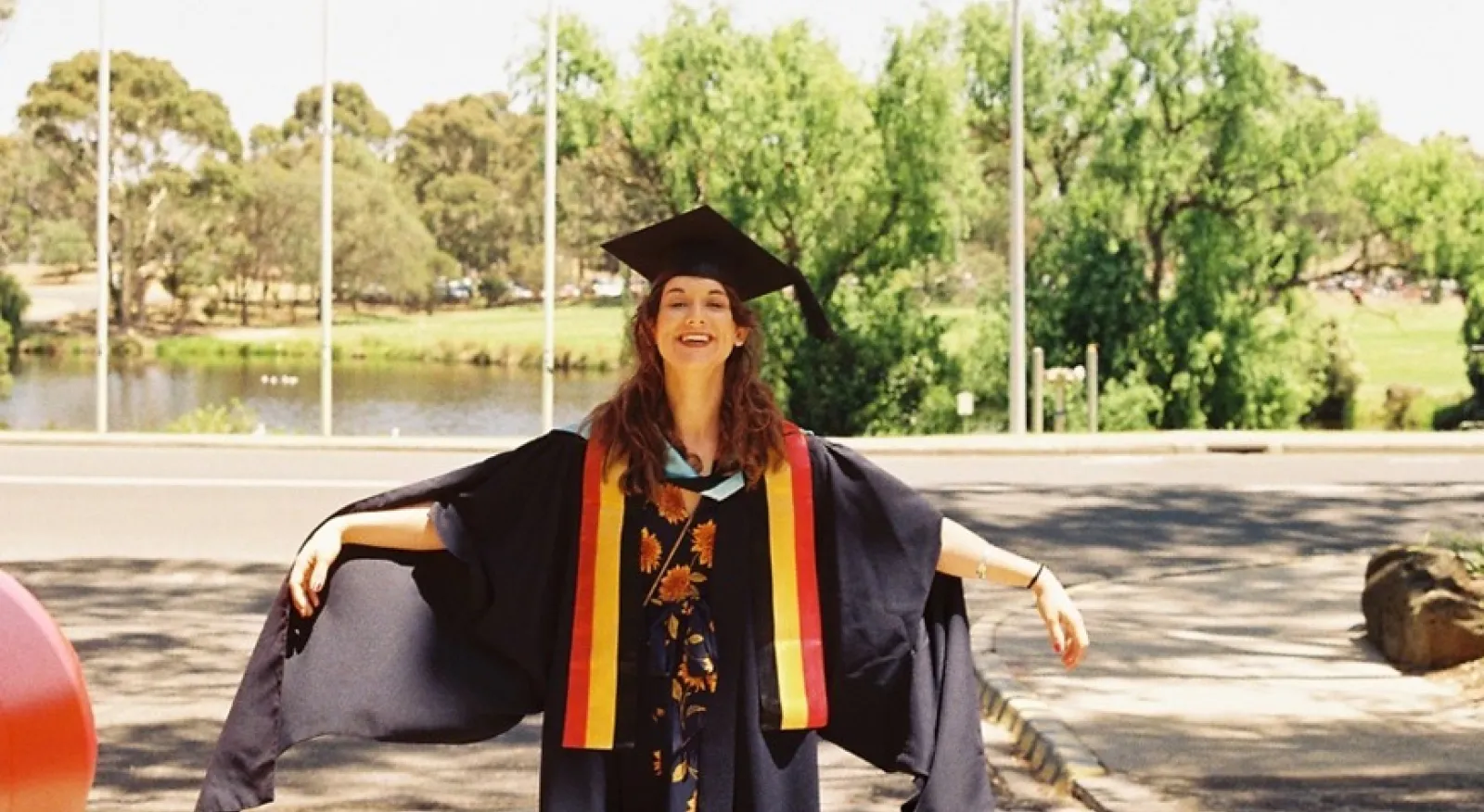 A woman in a graduation gown and cap proudly stands in front of a riverbank flanked with trees. She is smiling and holding her arms out.
