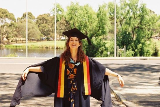 A woman in a graduation gown and cap proudly stands in front of a riverbank flanked with trees. She is smiling and holding her arms out.