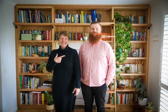 A man and a woman standing next to each other in front of a book case. The woman, wearing black, is pointing to the man next to her - a tall man with a beard and a pink shirt. They are both slightly smiling.