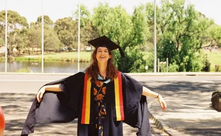 A woman in a graduation gown and cap proudly stands in front of a riverbank flanked with trees. She is smiling and holding her arms out.