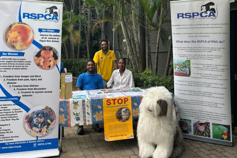 3 people at an exhibition booth promoting RSPCA PNG. There is a table, two banners and a promotional stuffed dog.