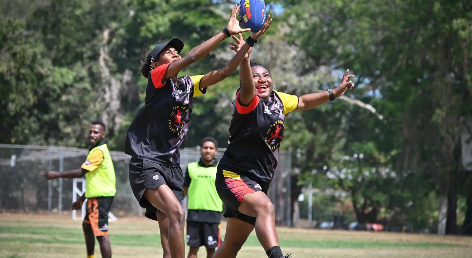Two people are playing AFL football and reaching for the ball mid-air. They're wearing team uniforms and smiling in an outdoor setting.