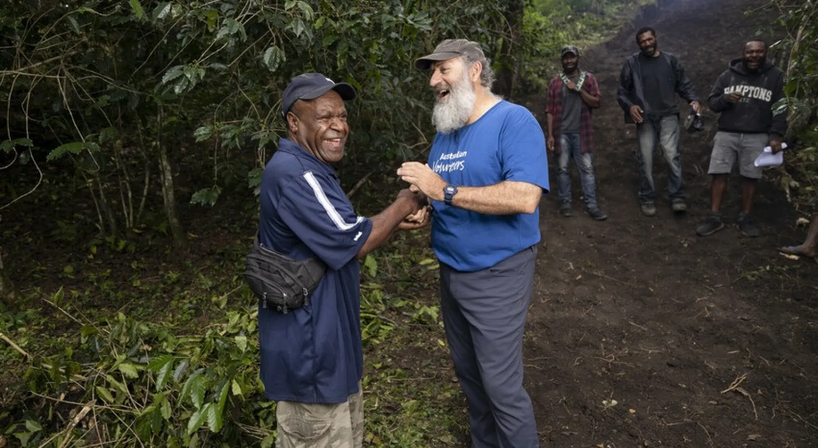 Two people are shaking hands and smiling broadly in a tropical outdoor setting