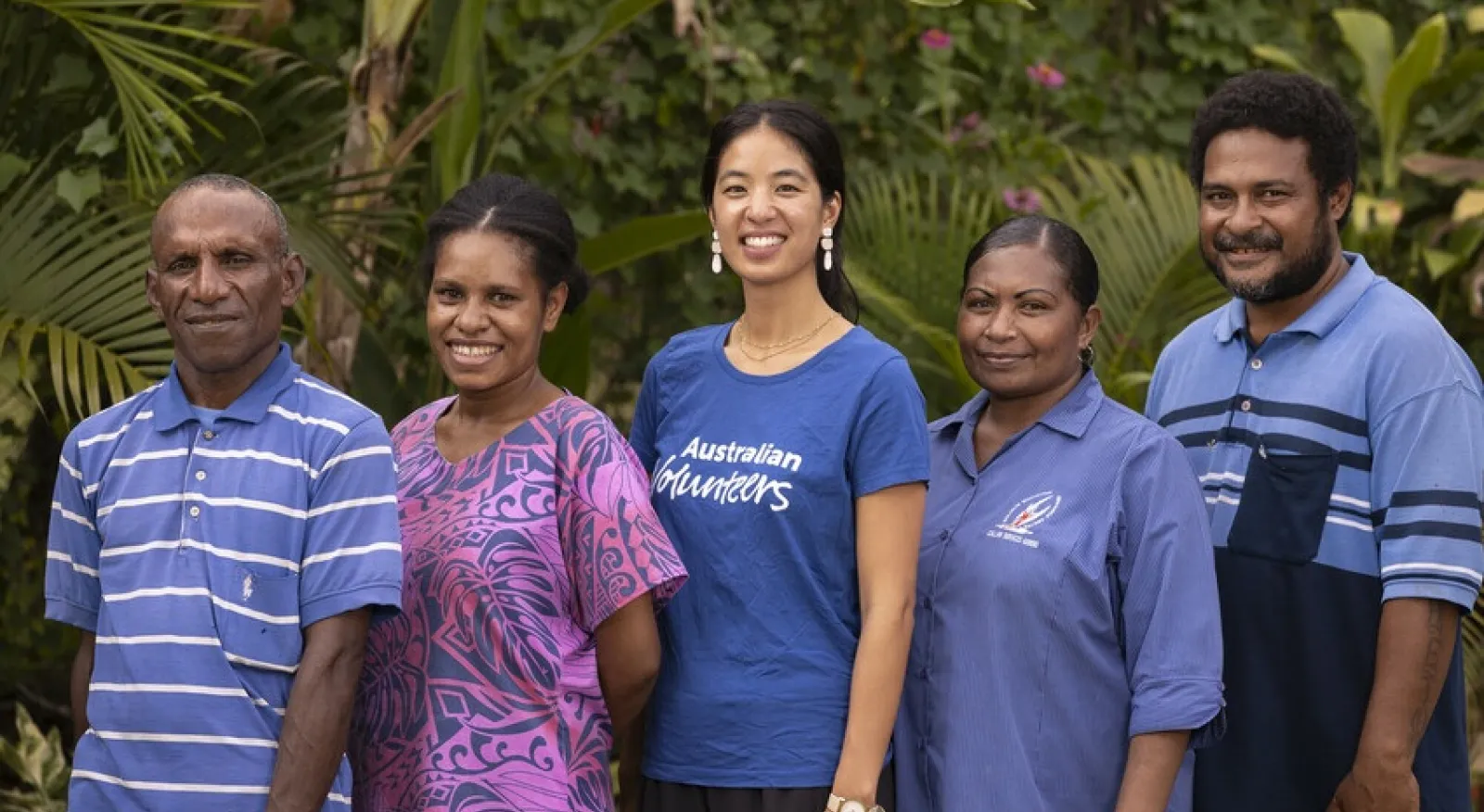 Five people stand in a line, angled towards - and smiling directly at - the camera. They are standing in front of greenery.