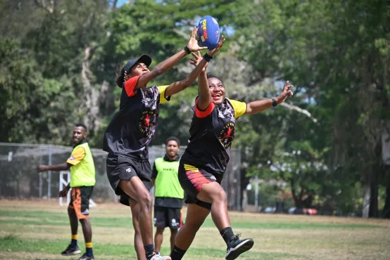 Two people are playing AFL football and reaching for the ball mid-air. They're wearing team uniforms and smiling in an outdoor setting.