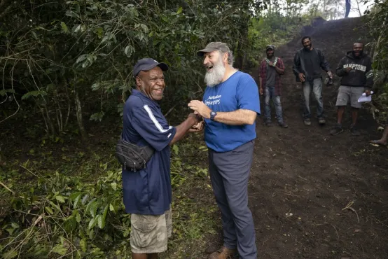 Two people are shaking hands and smiling broadly in a tropical outdoor setting