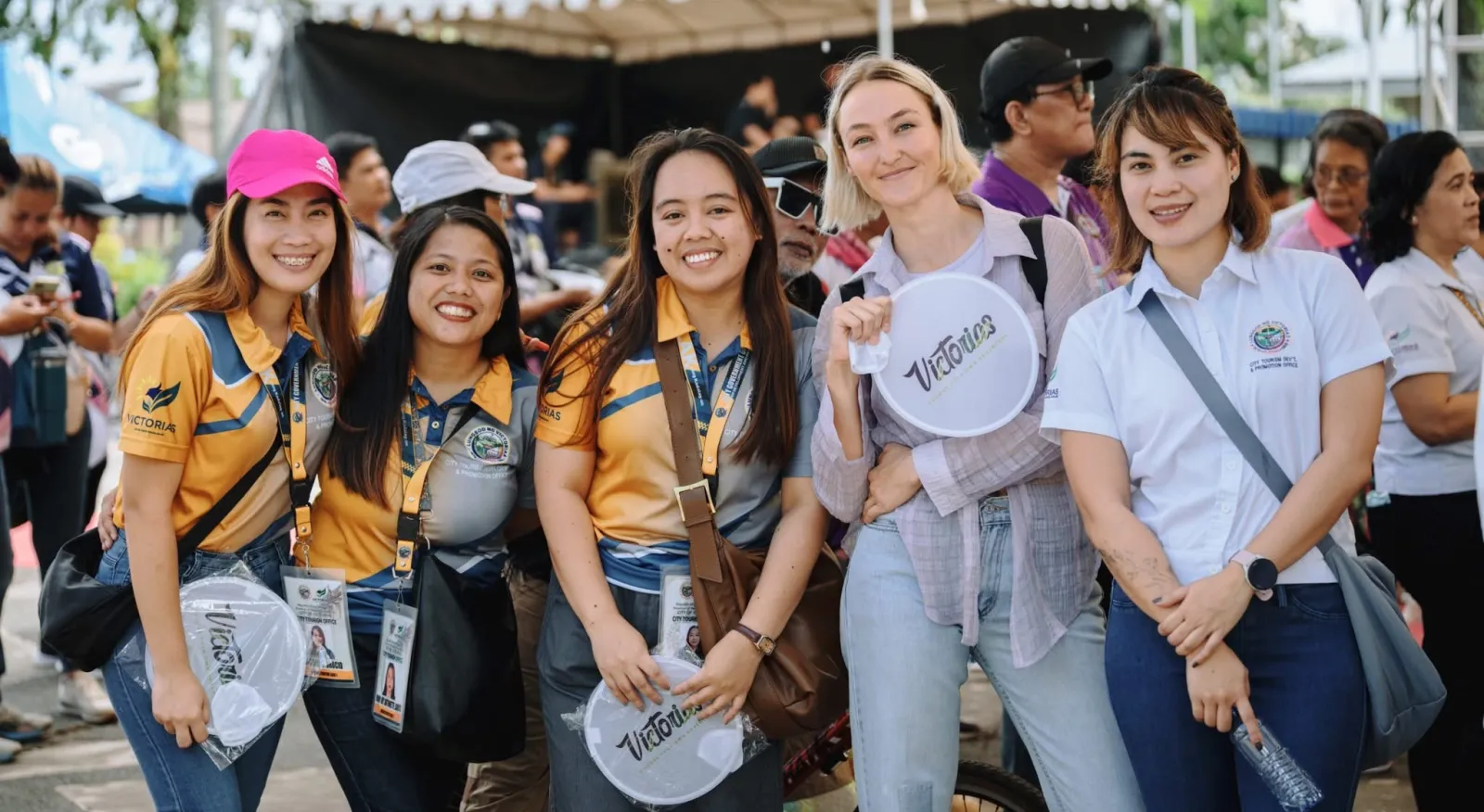 Five women pose and smile at the camera. All are in jeans and tshirts. They hold fans saying Victorias City