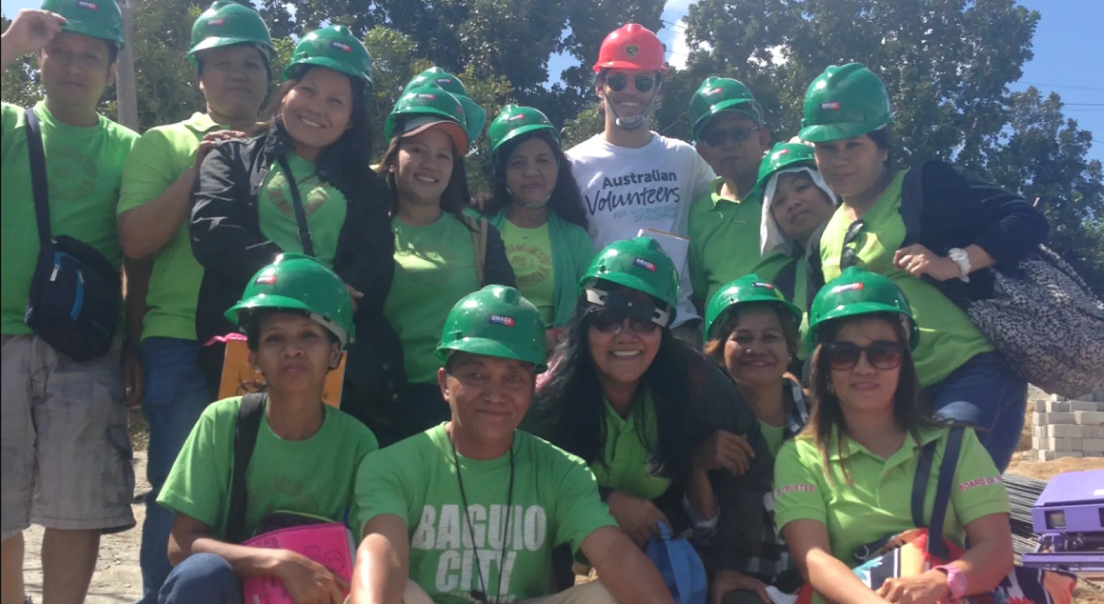 A group of people with green t-shirts wearing hard hats and smiling at the camera. One man has a white t-shirt and red hard hat.