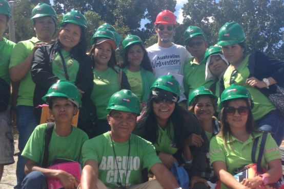 A group of people with green t-shirts wearing hard hats and smiling at the camera. One man has a white t-shirt and red hard hat.