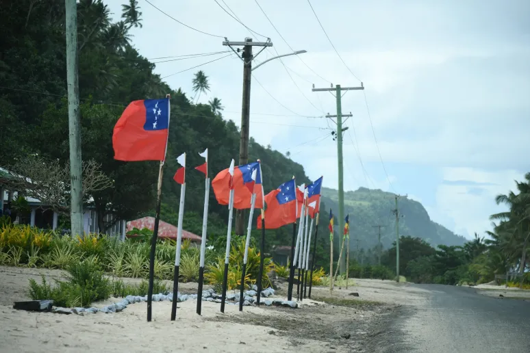 A row of flags lining a road, adding a vibrant touch to the surroundings of trees and and small mountains.
