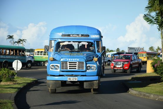 A blue Toyota van driving out of a bus depot, with cars and colourful mini buses behind it.