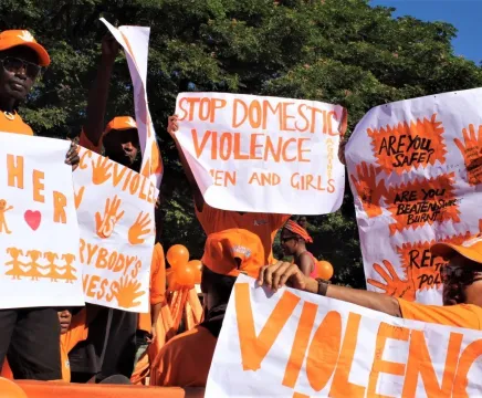Women and men hold multiple protest signs calling for the end of gender-based violence. Everyone is in orange, and the signs have orange writing as well.