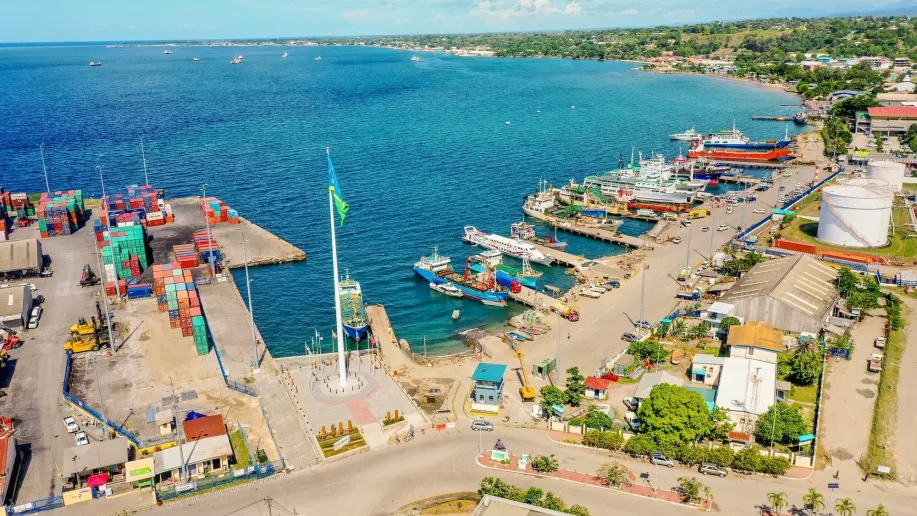 A busy port with crates and boats and a bright blue sea.