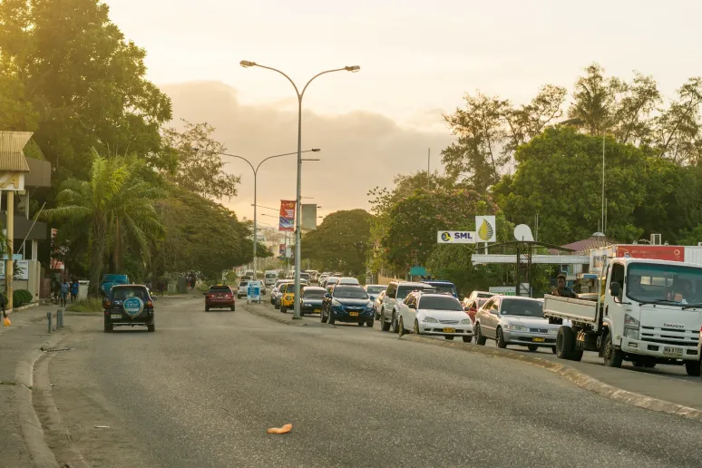 A view of a street approaching dusk, busy with cars and trucks, with green trees on either side of the roads.