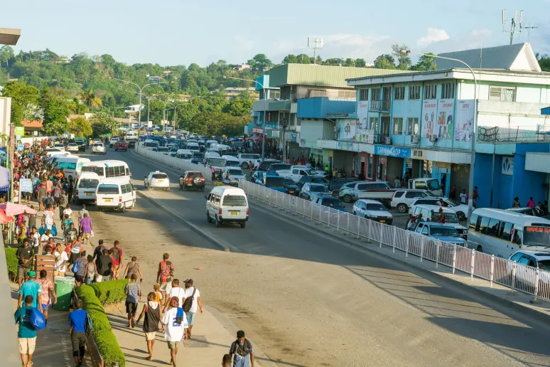 A bustling street with people on the sidewalk and cards and vans on the road.